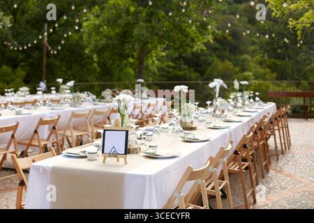 Ein wunderschön gestalteter Hochzeitsempfang im Freien verfügt über lange Tische mit weißen Tischdecken, Holzstühlen und zarte weiße Blumenarrangements un Stockfoto