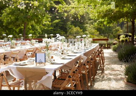 Elegante Hochzeitsatmosphäre im Freien mit langen Tischen mit weißen Blumenarrangements, rustikalen Holzstühlen und weichen Lichtern unter üppigen Lichtern Stockfoto