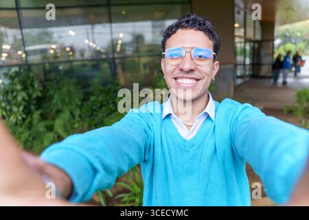 Ein junger hispanischer Mann aus lateinamerika, General Z, macht draußen ein Selfie mit einem fröhlichen Lächeln. Seine moderne Kleidung und sein fröhlicher Ausdruck spiegeln das Wesen des Betrügers wider Stockfoto