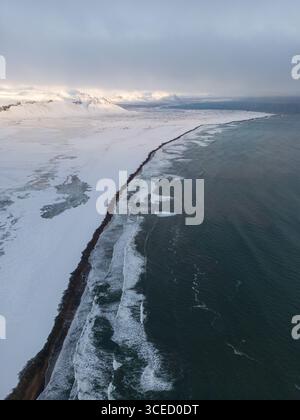Blick aus der Vogelperspektive auf eine isländische Winterlandschaft mit einer atemberaubenden Küste mit schneebedecktem Gelände und Wellen, die am Ufer abstürzen. Wild, REM Stockfoto