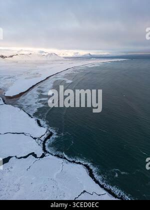 Blick aus der Vogelperspektive auf Islands verschneite Küste, wo zerklüftete Klippen auf den kalten Atlantik treffen. Die weitläufige Winterlandschaft fängt die heitere Schönheit ein Stockfoto