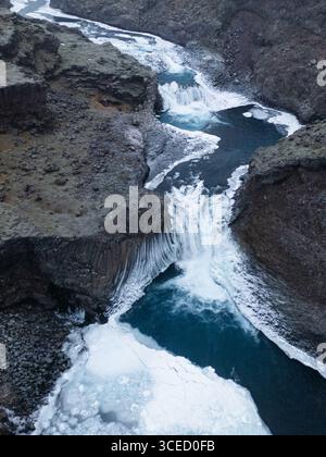 Aus der Vogelperspektive auf eine atemberaubende isländische Winterlandschaft mit einem majestätischen gefrorenen Wasserfall, der durch raues Gelände schneidet und die Wildnis hervorhebt Stockfoto