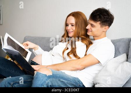 Ein junges Paar sitzt auf der Couch und erwartet freudig die Ankunft ihres Babys, während sie ein Fotoalbum anschauen, einen zärtlichen Moment der Liebe festhalten Stockfoto