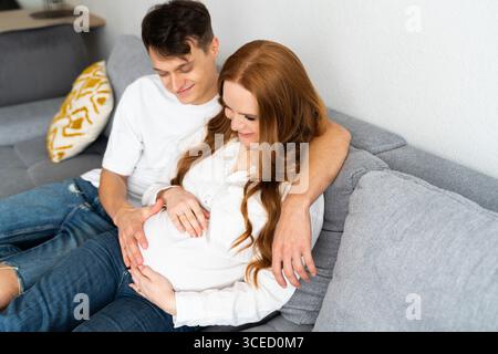 Ein junges Paar sitzt zusammen auf einer bequemen grauen Couch und erwartet sehnsüchtig ihre neue Ankunft. Die Hände der werdenden Mutter liegen liebevoll auf ihrem Ba Stockfoto