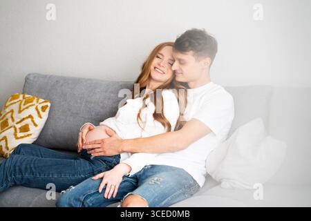 Ein junges Paar sitzt zusammen auf einer gemütlichen Couch und strahlt Glück und Liebe aus, während sie die Ankunft ihres Babys eifrig vorwegnehmen und ein zärtliches Gesicht hervorheben Stockfoto