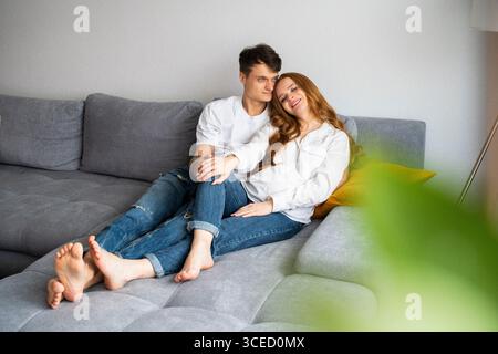 Ein junges Paar, das ein Baby erwartet, sitzt liebevoll auf einer gemütlichen grauen Couch, umhüllt und lächelt warm. Der zärtliche Moment hebt Liebe, Vorfreude hervor Stockfoto