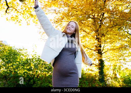 Schöne schwangere Frau mit rotem Haar posiert fröhlich in einer ruhigen Herbstkulisse, umgeben von lebendigem Herbstlaub, das die Essenz der Wärme einfängt Stockfoto