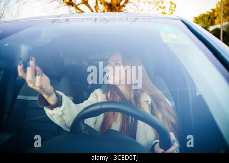 Eine schöne schwangere Frau mit langen roten Haaren lächelt, als sie im Auto sitzt und den Rückspiegel verstellt. Sonnenlichtfilter durch die Windschutzscheibe, Stockfoto