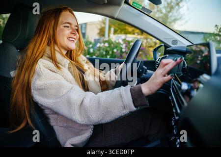 Eine schöne schwangere Frau mit langen roten Haaren fährt ein Auto und genießt einen sonnigen Tag. Ihr Lächeln strahlt Glück aus und zeigt einen aufmunternden Moment der Freiheit Stockfoto