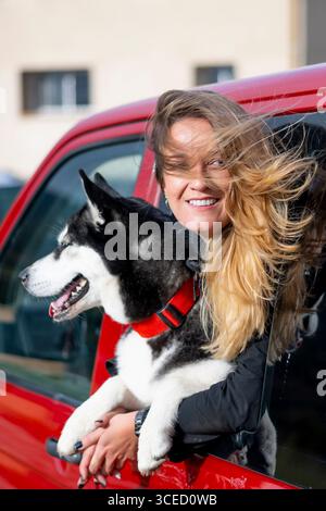 Eine lächelnde Frau mit ihrem Husky-Hund beugt sich vor Aufregung aus einem roten Auto. Die Zunge des Hundes ist raus, und ihr Haar weht im Wind und fängt eine Freude ein Stockfoto