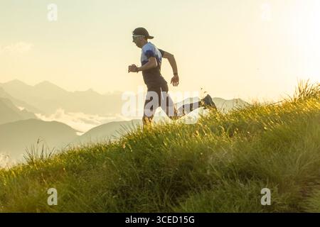 Ein Mann mit Mütze und Sonnenbrille läuft während des Sonnenuntergangs auf einem grasbewachsenen Hügel entlang, umgeben von einer Berglandschaft. Das Bild erfasst das Wesen von Stockfoto