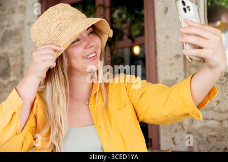 Eine junge, fröhliche Frau mit gelbem Hemd und Hut macht ein Selfie mit ihrem Smartphone, während sie allein an einem Café-Tisch sitzt und einen Moment von j festnimmt Stockfoto