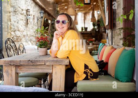 Eine junge, fröhliche Frau in einem gelben Hemd sitzt an einem Kaffeetisch und lächelt hell. Sie trägt eine Sonnenbrille und verleiht ihr einen Hauch von Stil Stockfoto
