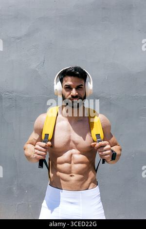 Muskulöser Mann mit Kopfhörern und gelbem Rucksack steht selbstbewusst vor einer strukturierten grauen Wand und fängt die Essenz von Fitness und modernem Lifest ein Stockfoto