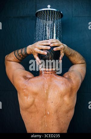 Ein Mann mit Tätowierungen genießt eine erfrischende Dusche und steht mit dem Rücken zur Kamera. Das Wasser fällt aus dem Duschkopf und sorgt für eine entspannende und entspannende Atmosphäre Stockfoto