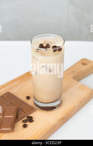 Ein erfrischender Eiskaffee in einem hohen Glas mit Kaffeebohnen auf einem Holzbrett mit Schokoladenstückchen. Das ideale Sommergetränk für Kaffeeliebhaber Stockfoto