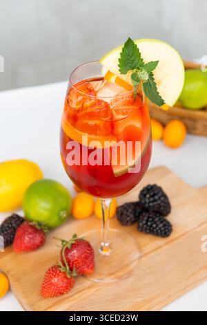Erfrischendes Sommergetränk in einem Glas mit Eis, garniert mit Apfel und Minze. Umgeben von frischen Früchten wie Erdbeeren, Brombeeren und Zitrusfrüchten Stockfoto