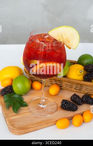 Ein lebendiges Glas Sangria gefüllt mit Eis, Zitrusscheiben und einem Stück Apfel darauf. Das Getränk wird auf einem Holzbrett platziert, umgeben von frischem Berr Stockfoto