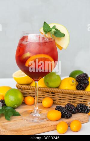 Ein Glas roter Sangria gefüllt mit Zitrusscheiben und Minze sitzt auf einem Holzbrett. Rund um das Getränk gibt es eine Auswahl an Brombeeren und Zitrusfrüchten Stockfoto