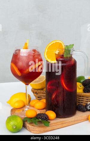 Eine erfrischende Sommerszene mit einem Glas und Krug aus roter Sangria mit Zitrusscheiben und Beeren, umgeben von frischem Obst auf einem Holztablett aga Stockfoto