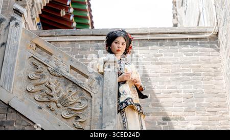 Eine Frau in traditioneller Kleidung der Qing-Dynastie steht auf einer kunstvollen Steintreppe in Pingyao, China, und blickt direkt in die Kamera und reflektiert Stockfoto