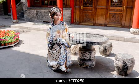 Eine Frau in Kleidung der Qing-Dynastie sitzt draußen und hält einen dekorativen Ventilator in Pingyao, China. Sie ist umgeben von traditioneller Architektur A Stockfoto