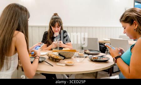 Drei junge Frauen sitzen an einem stilvollen Restauranttisch in ihre Smartphones. Der Tisch ist mit einer Vielzahl von Gerichten besetzt, die kreieren Stockfoto