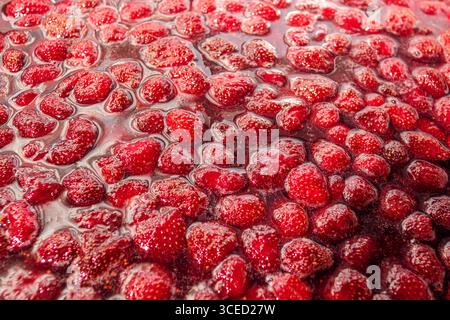 Eine Nahaufnahme hausgemachter Erdbeermarmelade mit leuchtender roter Farbe und glitzernder Textur. Die Marmelade ist mit ganzen Erdbeeren gefüllt, Reflec Stockfoto