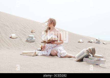 Eine Frau in einem weißen Kleid sitzt auf dem Sand, umgeben von offenen und fliegenden Büchern, und schafft eine surreale und fantasievolle Szene des Friedens, der Kreativität und des Fans Stockfoto