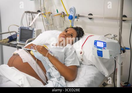 Eine schwangere Frau liegt in einem Krankenhausbett und bereitet sich auf die Geburt vor. Umgeben von medizinischer Ausrüstung wartet sie mit Vorfreude und Hoffnung und verkörpert das Stockfoto