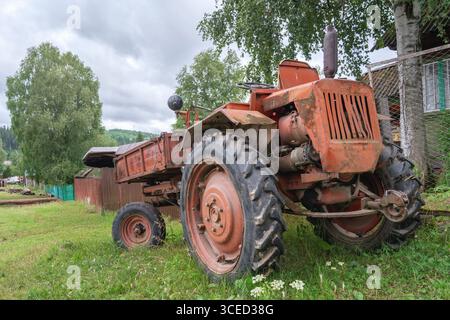 Der alte alte alte Traktor t16, der an einem bewölkten Tag auf dem Land in einem üppigen grünen Feld in der Nähe eines rustikalen Bauernhauses geparkt wurde. Stockfoto