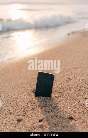 Smartphone steht bei Sonnenuntergang im Sand am Strand, Konzept eines Unfalls, Verlust, vergessenes oder beschädigtes elektronisches Gerät auf dem Seeweg. Selektiver Fokus. Stockfoto