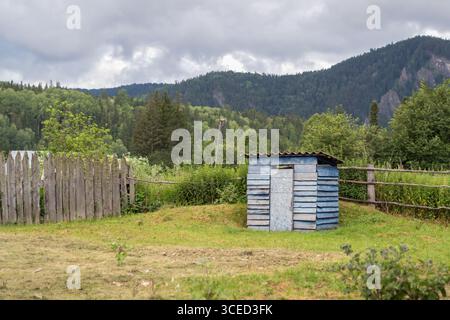 Rustikales altes altes Outdoor WC blaues Haus auf dem Hintergrund der wunderschönen Natur und Berge. Ein kleiner rustikaler Schuppen steht vor einem Hintergrund grüner Wälder Stockfoto