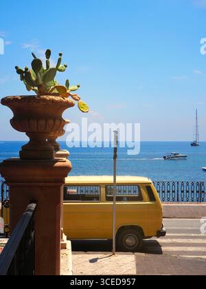 Gelber Vintage-Van, geparkt am Meer in Giardini Naxos in der Nähe von Taormina mit Kaktuspflanze und Blick auf das Mittelmeer unter hellem Sommerhimmel Stockfoto