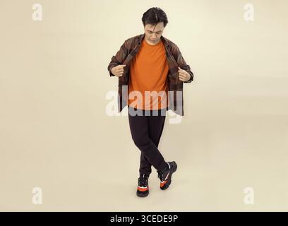 Ein Mann, der ein orangefarbenes Hemd und ein kariertes Überhemd trägt, posiert lässig vor einem beigefarbenen Hintergrund Stockfoto