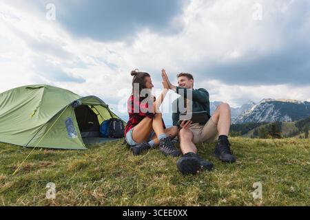 Zwei Camper teilen sich einen freudigen Moment in den Bergen, während sie auf dem Gras sitzen und von einem Zelt umgeben sind. Stockfoto