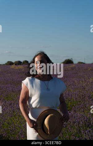 Eine Frau mit weißem Kleid und Strohhut steht auf einem Feld aus violetten Blumen. Sie lächelt und blickt in die Kamera Stockfoto