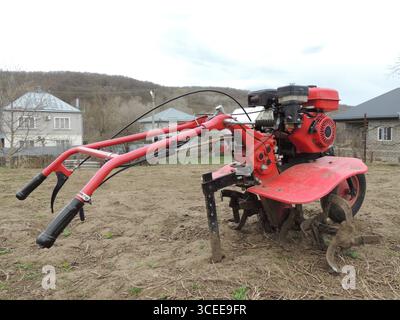 Roter Hand-Benzinpflug, der auf Bauernhof mit trockenem Gras vor dem Hintergrund von Dorfhäusern steht, Mechanisierung der privaten Landwirtschaft Stockfoto