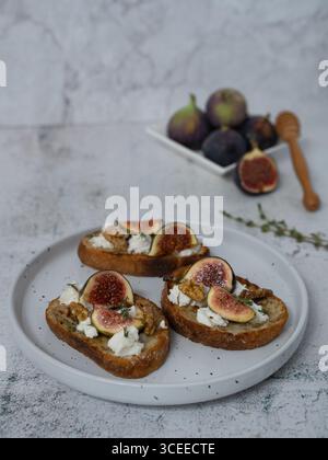 Vertikale Nahansicht von vorne von drei Feigen und Ziegenkäse auf weißer Platte mit Thymian, Gabel, grauem Hintergrund, Feigen auf separater Platte, Toast in b Stockfoto