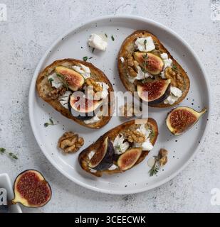 Horizontale Draufsicht Nahaufnahme von drei Feigen- und Ziegenkäse-Toasts auf weißer Platte mit Thymian, Feigen auf separatem Teller, herzhafte süße Mischung Stockfoto