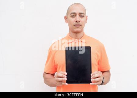 Studio-Porträt des gutaussehenden mittelalten hispanischen Mannes in orangefarbenem Poloshirt vor weißem Hintergrund mit digitalem Tablet-Computer. Kasuistik Stockfoto