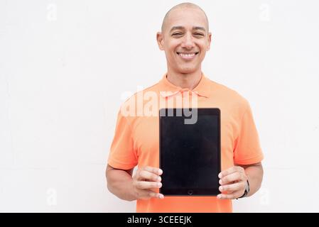 Studio-Porträt des gutaussehenden mittelalten hispanischen Mannes in orangefarbenem Poloshirt vor weißem Hintergrund mit digitalem Tablet-Computer. Kasuistik Stockfoto