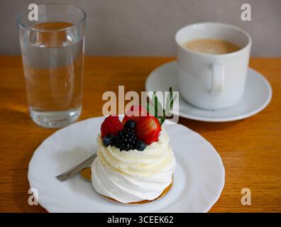 Kleiner pavlova-Kuchen mit Schlagsahne und frischen Beeren, serviert auf weißem Porzellanteller mit einem Glas Wasser und einer Kaffeetasse auf Holztisch Stockfoto