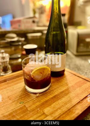Ein dunkler Cocktail, der in einem Steinglas mit Eis und Zitronengeschmack auf einem Holzbrett serviert wird, mit einer Flasche und Barausstattung im Hintergrund. Stockfoto