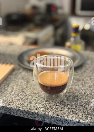 Ein klares Glas mit cremigem Eiskaffee steht auf einem Metalltisch, mit einem verschwommenen Sitzbereich im Hintergrund. Stockfoto
