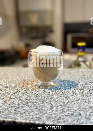Ein schäumendes Kaffeegetränk, das in einem klaren Glas mit einer Schaumschicht auf einem Kaffeetisch mit verschwommenem Hintergrund serviert wird. Stockfoto