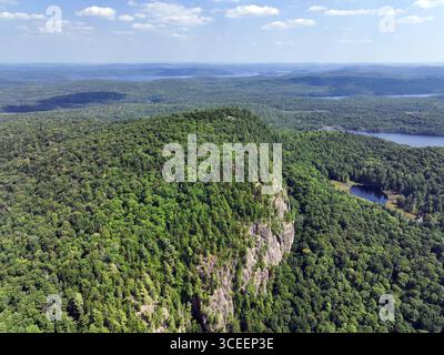 Steile, felsige Klippen ragen aus einer dicht bewaldeten Landschaft mit Seen und sanften Hügeln in der Ferne unter blauem Himmel hervor Stockfoto