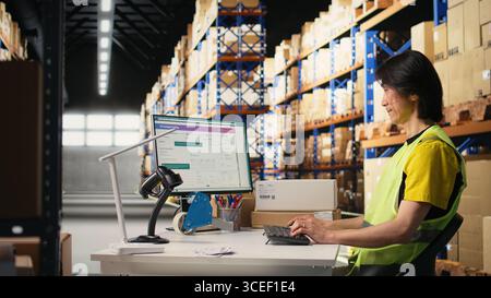 Zwei Kollegen arbeiten an der Auftragsbearbeitung in industriellen Lagerhäusern und tragen Warnwesten in der Nähe von riesigen Lagerregalen. Asiatischer Mann, der die Versanddaten auf dem Computer eingibt. Kamera A.. Stockfoto