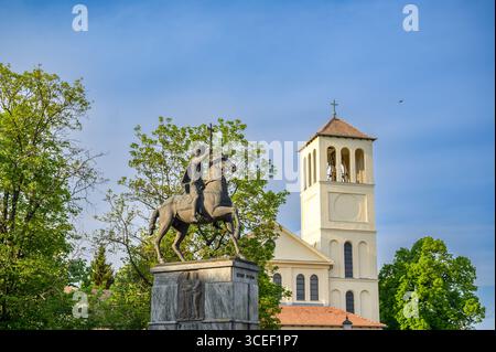 HODMEZOVASARHELY, UNGARN 06. MAI 2022 Eine bronzene Reiterstatue steht vor einer cremefarbenen Kirche unter klarem blauen Himmel. Üppig grüne Bäume Stockfoto