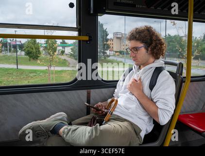 Prag, Tschechische Republik, 6. August 2023. Ein junger Kaukasier mit lockigen Haaren und Brille sitzt am Busbahnhof. Er wartet auf den Bus Stockfoto